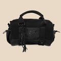 A black duffel bag with a top carry handle, adjustable shoulder strap, and braided tassel detail, shown against a solid beige background.