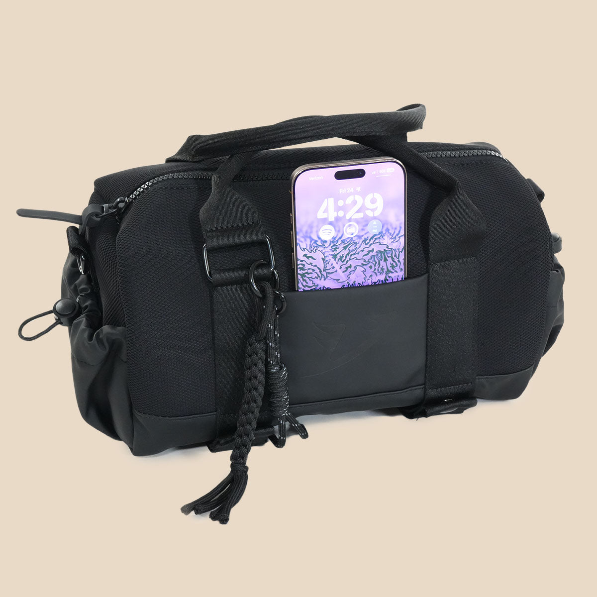 A black tactical-style duffel bag with a top handle, side zipper, braided hanging strap, and a smartphone inserted into a front pocket, shown on a plain beige background.