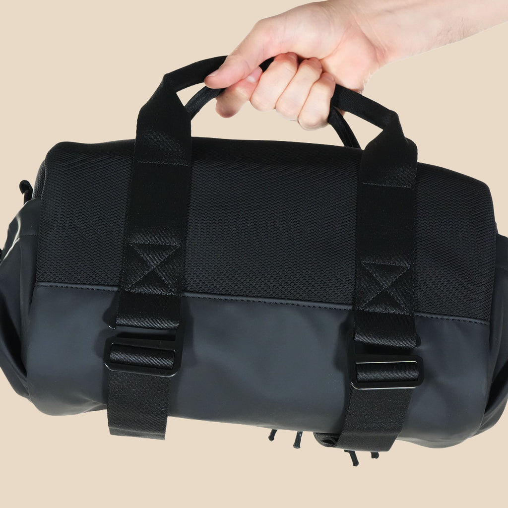 Orion black duffel bag with dual top handles and adjustable webbing straps, held by a person's hand against a solid beige background.