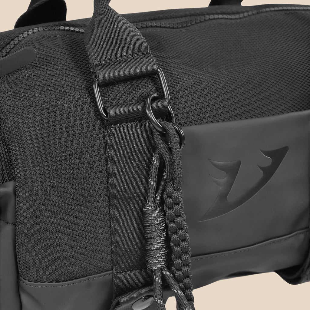 Close-up of a black mini duffel with a textured mesh fabric, a wide adjustable webbing strap with a metal buckle, a braided black cord keychain, and a matte black front  panel featuring an embossed Vantagym logo.
